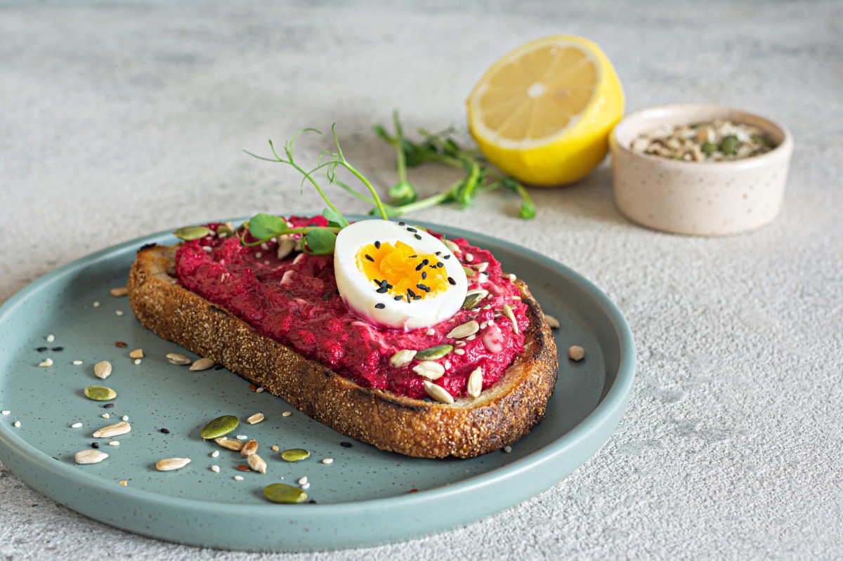 Eine Scheibe Brot mit Rote-Bete-Aufstrich mit FrischkÃ¤se und Ei auf einem Teller, garniert mit Kernen, im Hintergrund eine halbe Zitrone und eine Schale Kerne.