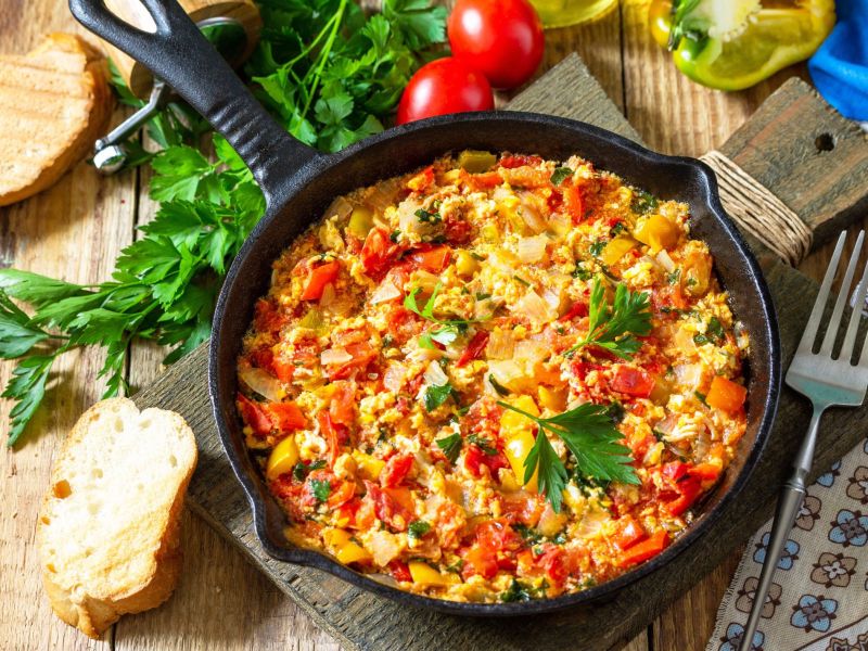 RÃ¼hrei mit GemÃ¼se, serviert in einer gusseisernen Pfanne. Im Hintergrund liegen KrÃ¤uter, kleine Tomaten und frisches Brot als Deko.