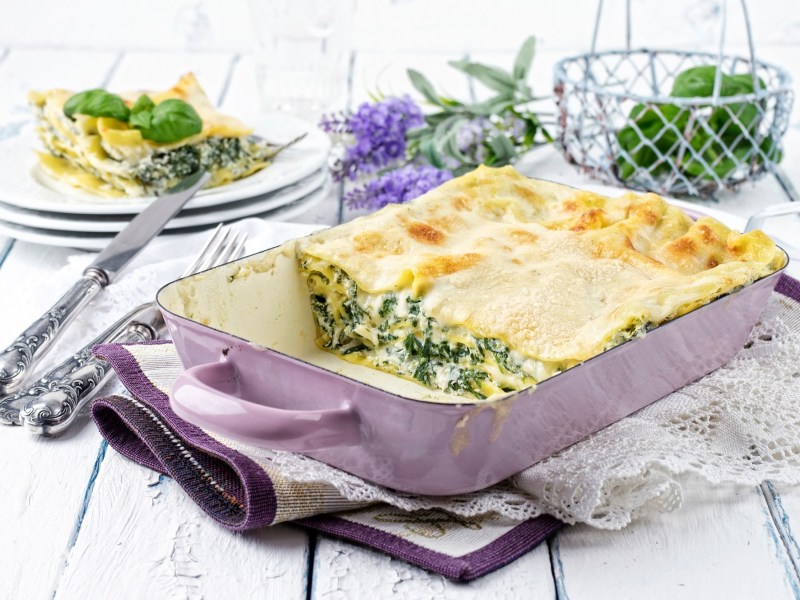 Spinat-Ricotta-Lasagne in einer Auflaufform, ein StÃ¼ck bereits herausgenommen und auf einem Teller im Hintergrund.