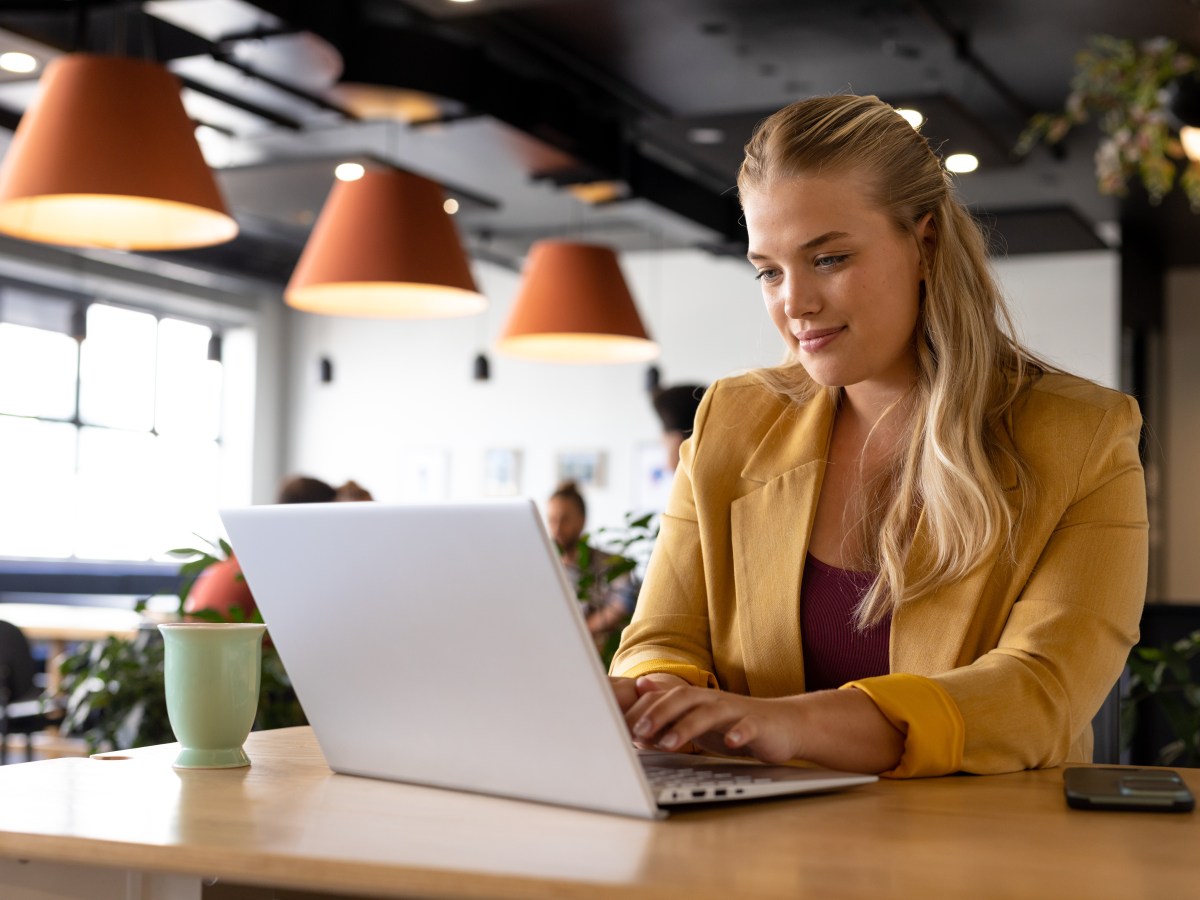 Eine Frau mit blonden Haaren sitzt im BÃ¼ro und arbeitet am Laptop.