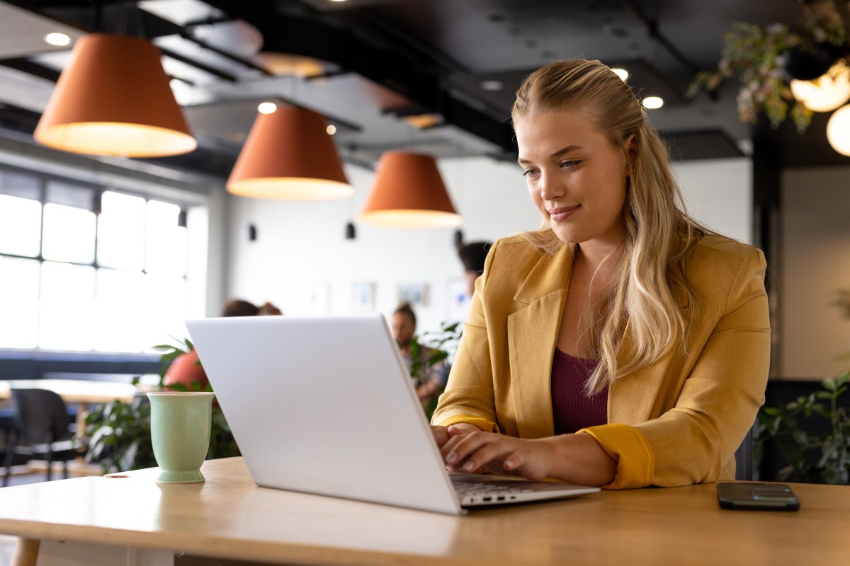 Eine Frau mit blonden Haaren sitzt im BÃ¼ro und arbeitet am Laptop.