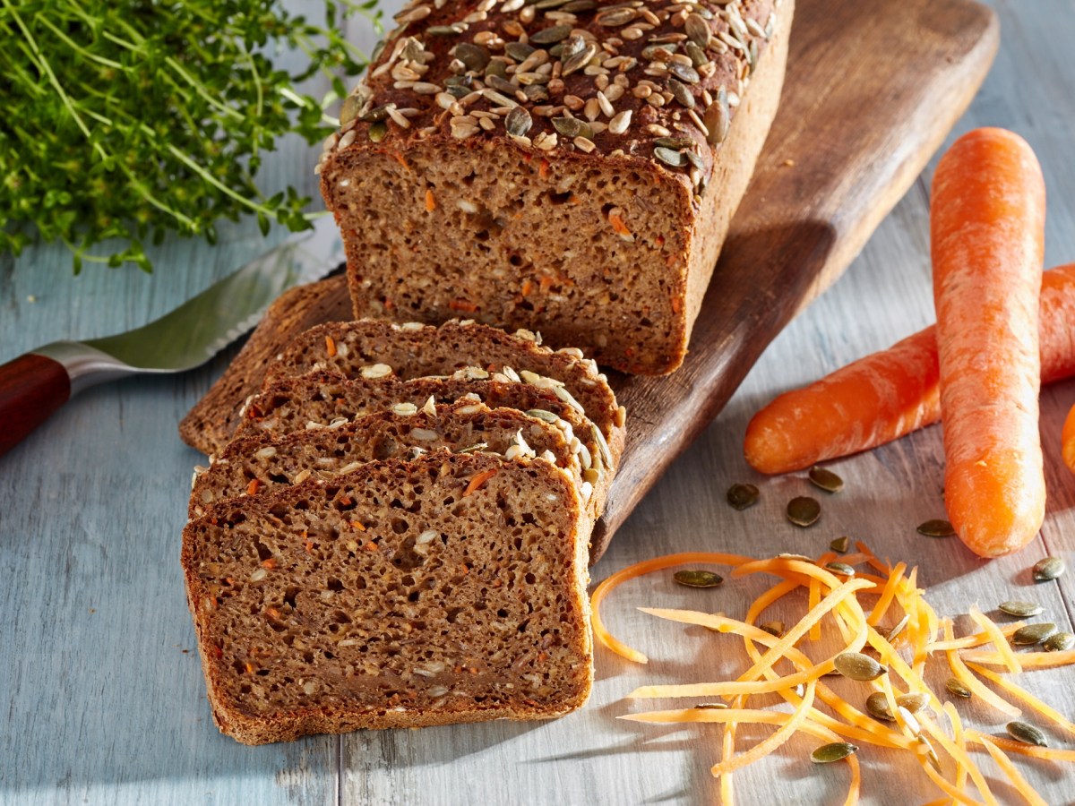 Vollkornbrot mit MÃ¶hren, angeschnitten, auf einem Holzbrett. Daneben liegen MÃ¶hren.