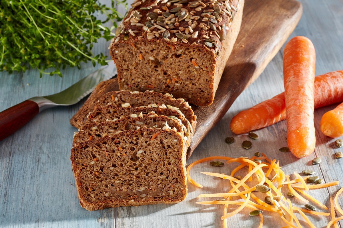 Vollkornbrot mit MÃ¶hren, angeschnitten, auf einem Holzbrett. Daneben liegen MÃ¶hren.