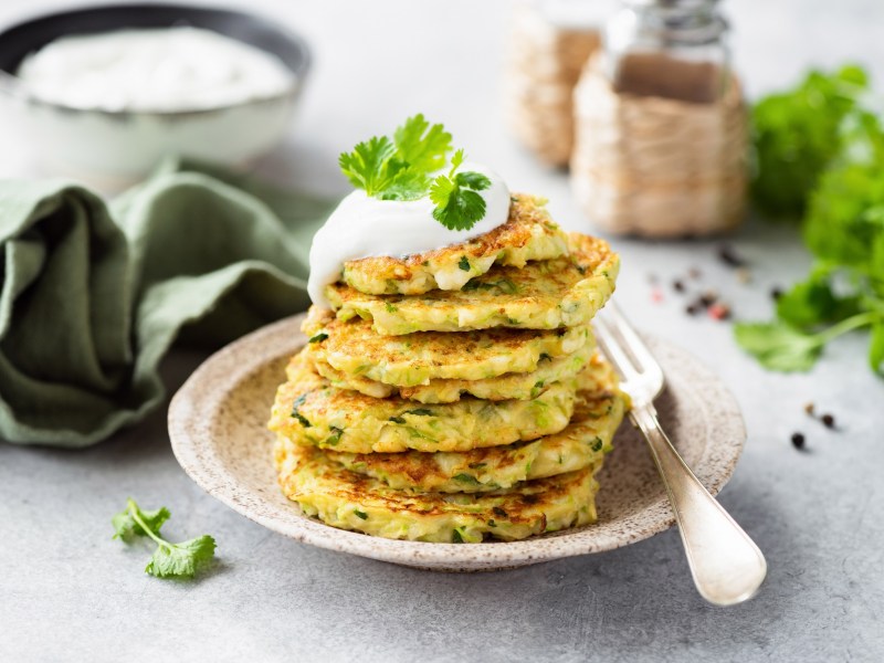 Ein Stapel Zucchini-Pancakes mit HÃ¼ttenkÃ¤se auf einem weiÃŸen Teller. Daneben liegt eine Gabel. Die Pancakes sind mit Joghurt und Petersilie garniert.