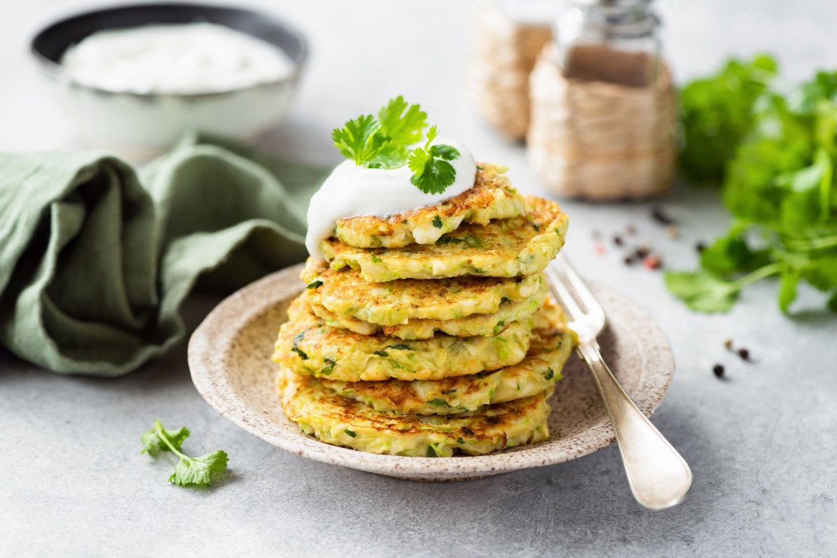 Ein Stapel Zucchini-Pancakes mit HÃ¼ttenkÃ¤se auf einem weiÃŸen Teller. Daneben liegt eine Gabel. Die Pancakes sind mit Joghurt und Petersilie garniert.