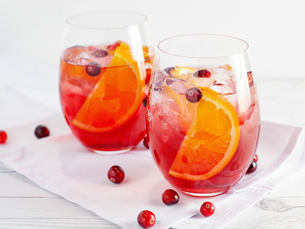 Zwei GlÃ¤ser mit Cranberry-Orangen-Spritz stehen auf einer weiÃŸen Serviette. Als Dekoration liegen einige Cranberrys neben den GlÃ¤sern.