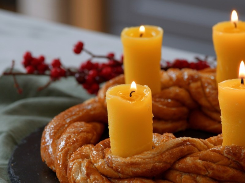 Ein essbarer Adventskranz aus Brezelteig mit vier Kerzen aus Butter. Im Hintergrund eine grÃ¼ne Serviette und rote Pflanzen, passend zu Weihnachten.