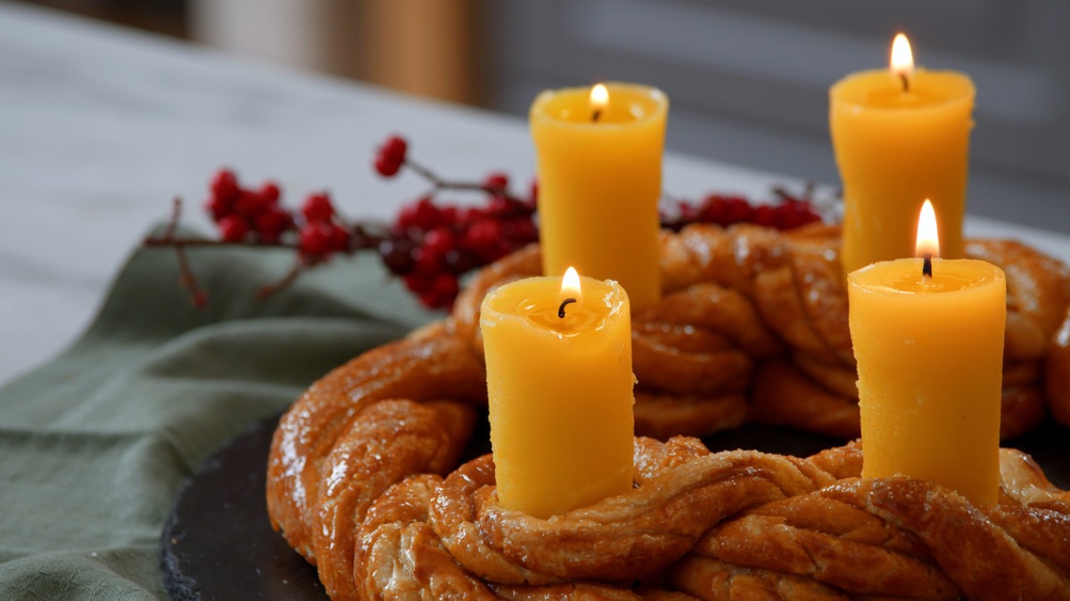 Ein essbarer Adventskranz aus Brezelteig mit vier Kerzen aus Butter. Im Hintergrund eine grÃ¼ne Serviette und rote Pflanzen, passend zu Weihnachten.