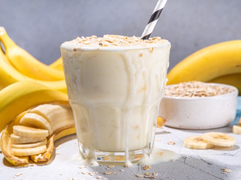 Ein Glas FrÃ¼hstÃ¼cksshake mit Bananen und Haferflocken. Im Hintergrund sind Bananen uds eine Schale mit Haferflocke zu sehen.