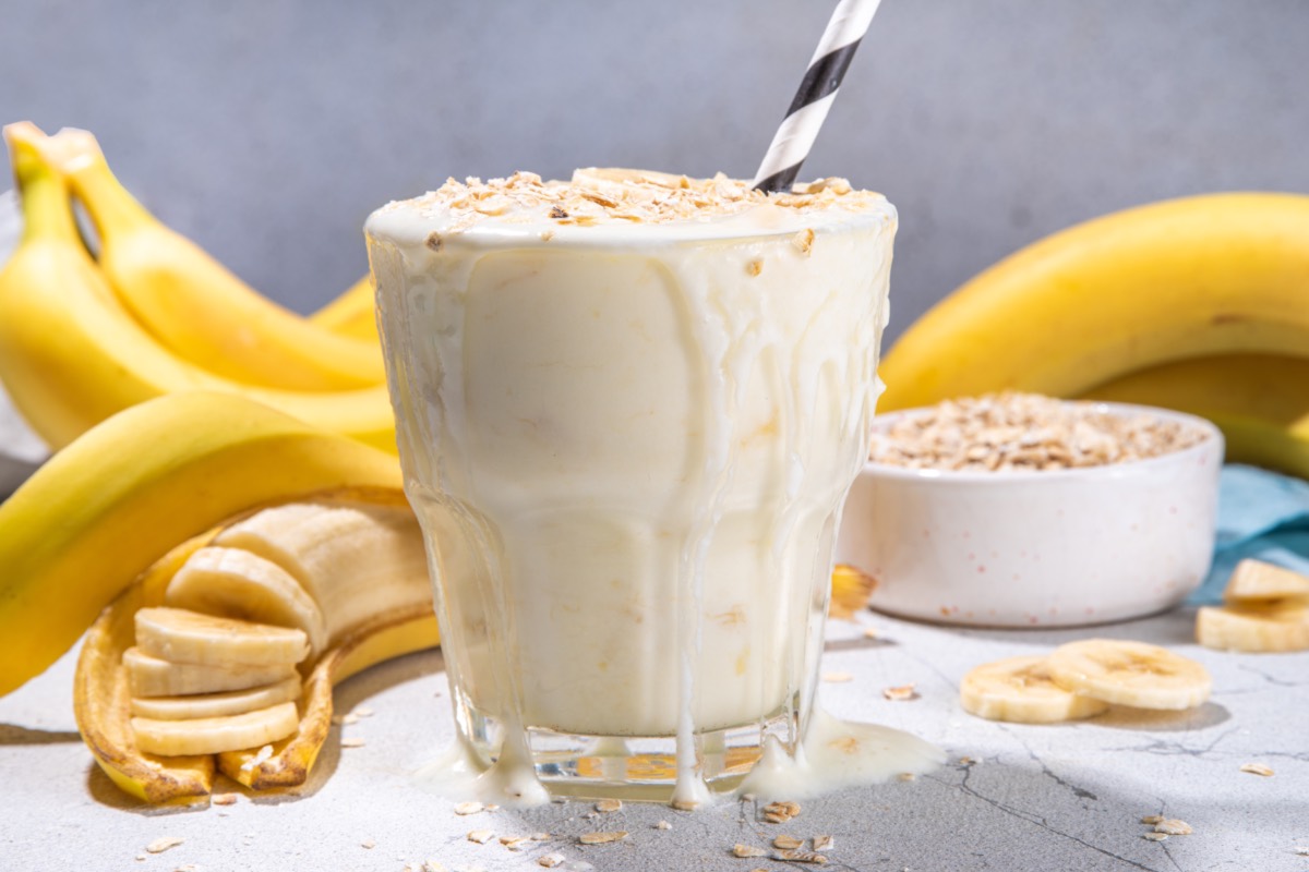 Ein Glas FrÃ¼hstÃ¼cksshake mit Bananen und Haferflocken. Im Hintergrund sind Bananen uds eine Schale mit Haferflocke zu sehen.