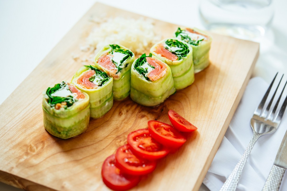 6 Gurken-RÃ¶llchen mit Lachs und Feta auf einem Brett, daneben in Scheiben geschnittene Tomaten.