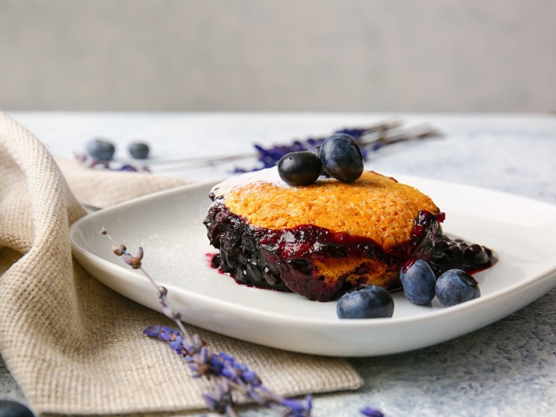 Eine Portion Heidelbeer-Auflauf mit frischen Blaubeeren auf einem Teller, daneben drapiert eine Stoffserviette und BlÃ¼ten.