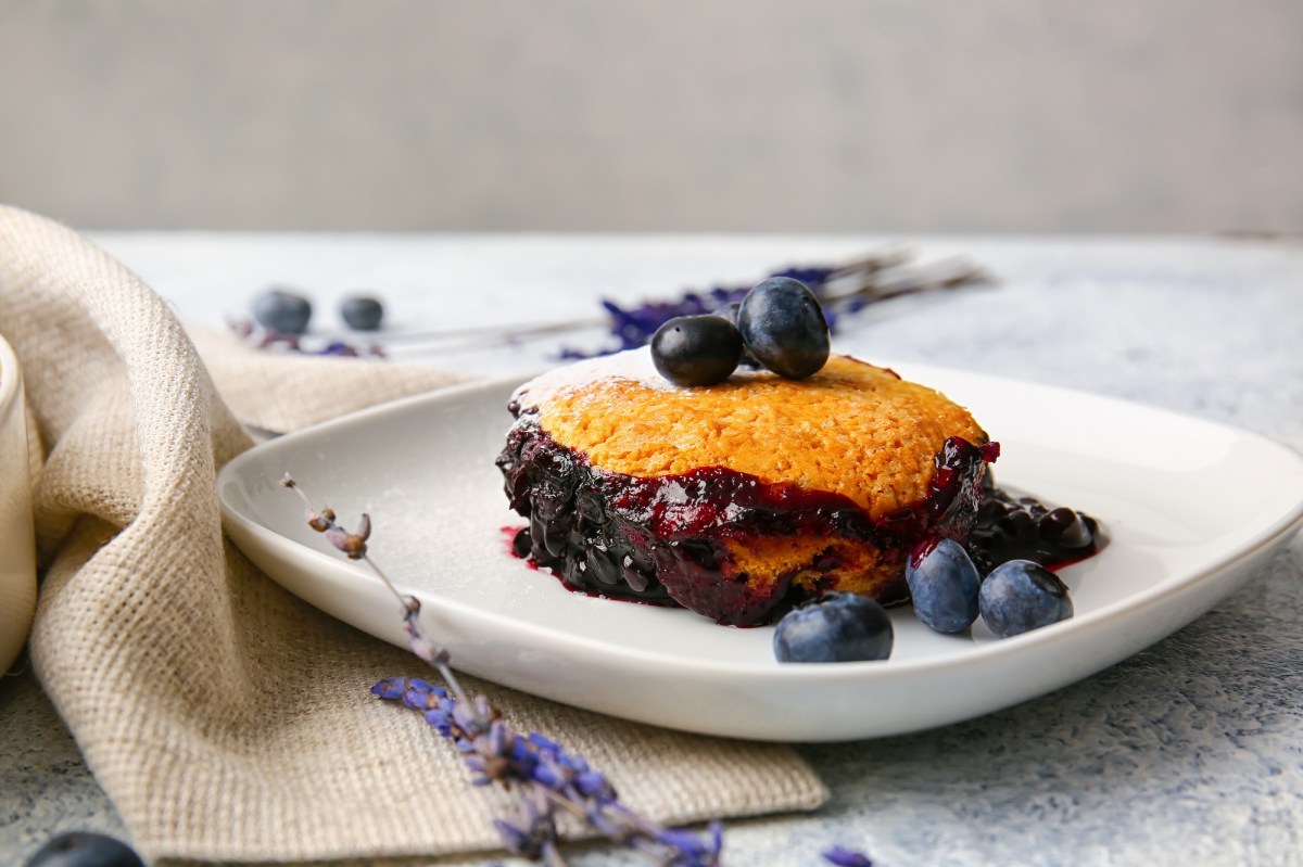 Eine Portion Heidelbeer-Auflauf mit frischen Blaubeeren auf einem Teller, daneben drapiert eine Stoffserviette und BlÃ¼ten.