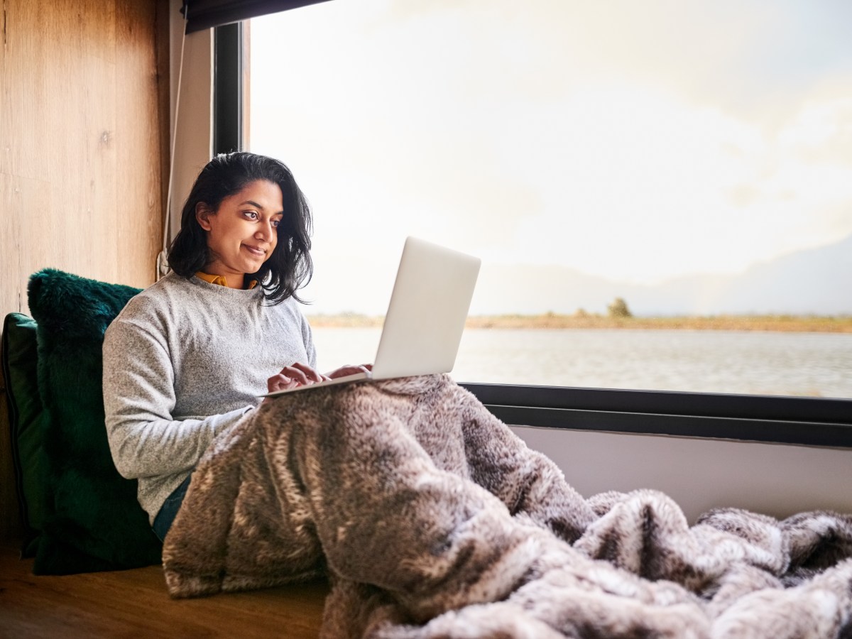 Frau sitzt mit Decke am groÃŸen Fenster und benutzt Laptop zu Hause.