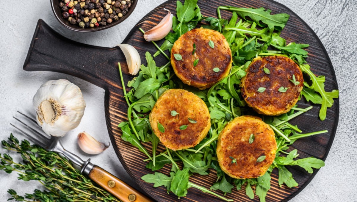 Vier Linsenfrikadellen auf einem Bett aus Rucola auf einem runden Holzbrett. Daneben steht eine Schale mit Pfeffer und es liegen Knoblauchzehen herum.