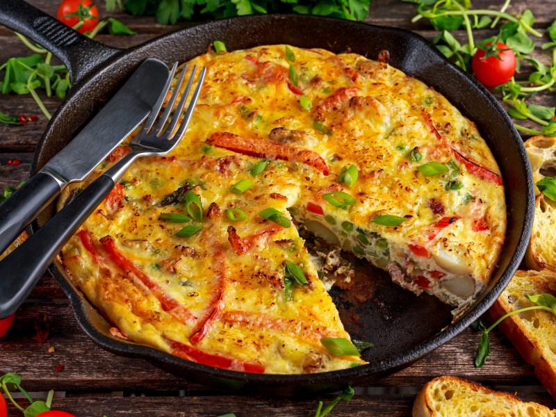 Quiche-Auflauf, von dem ein StÃ¼ck fehlt, in einer gusseisernen Pfanne. Darauf liegen Messer und Gabel und drumherum ist der Tisch mit Brot und Rucola bedeckt.