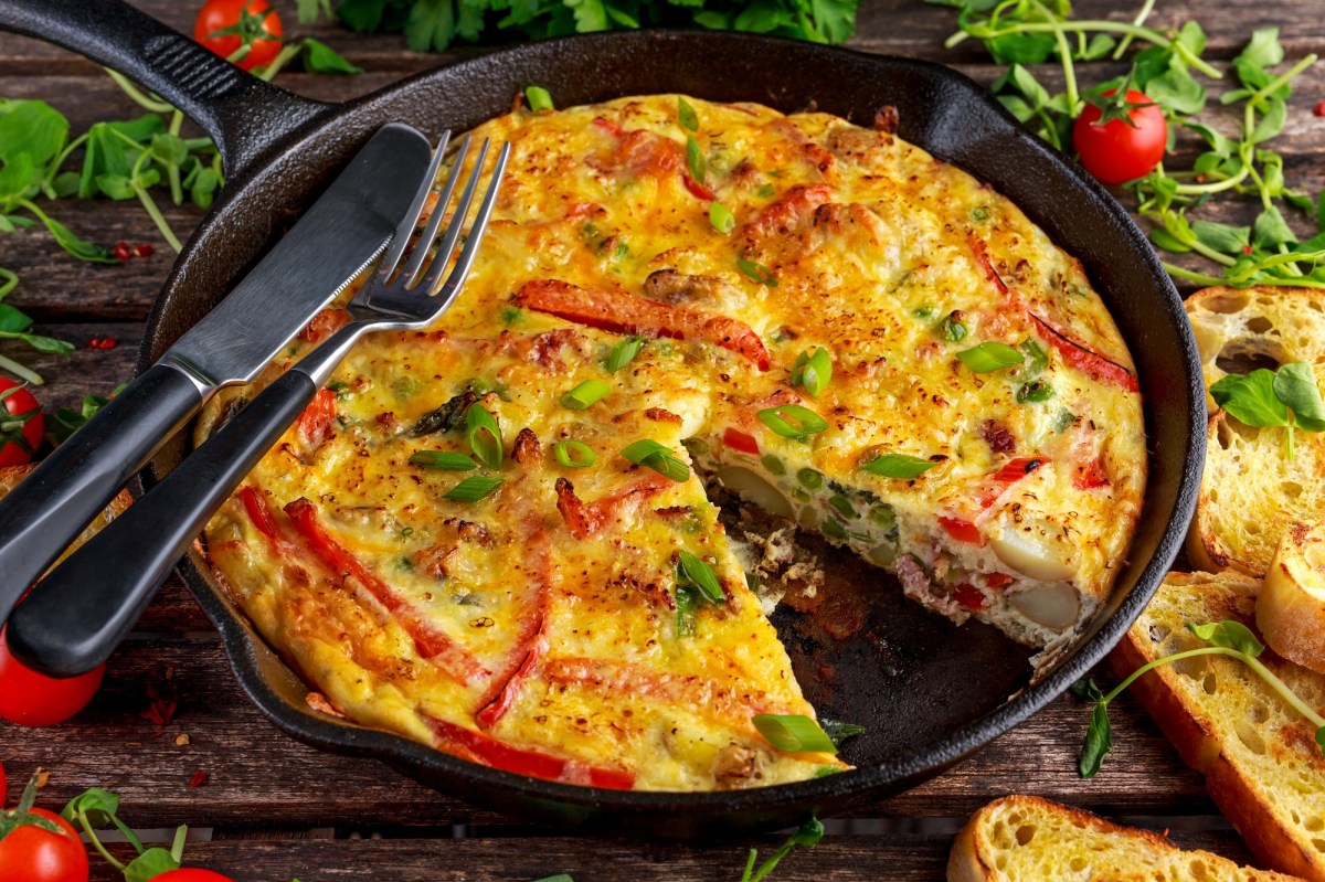 Quiche-Auflauf, von dem ein StÃ¼ck fehlt, in einer gusseisernen Pfanne. Darauf liegen Messer und Gabel und drumherum ist der Tisch mit Brot und Rucola bedeckt.