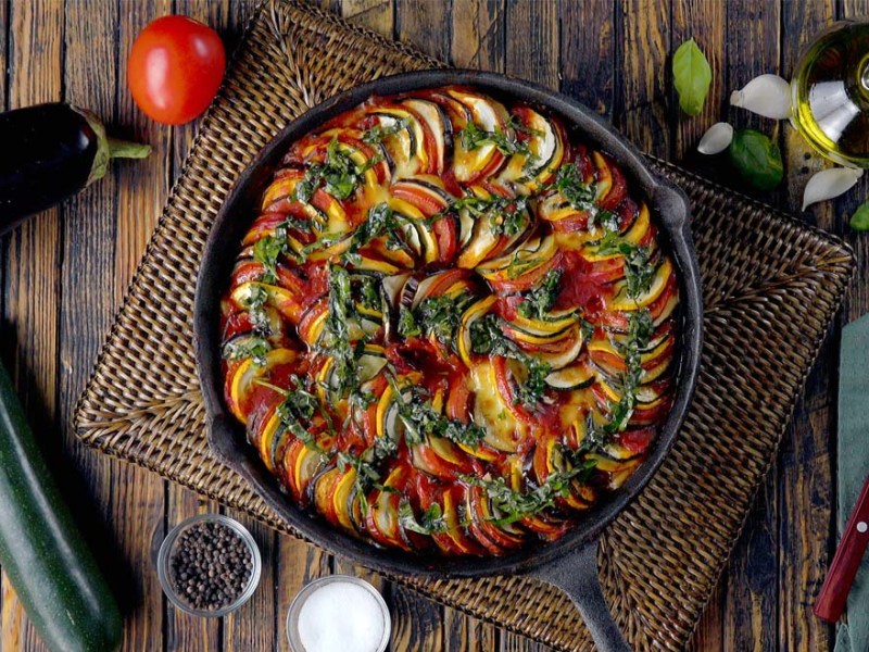 Ratatouille in der Pfanne, von oben fotografiert. Daneben liegen Besteck und frische Zutaten.