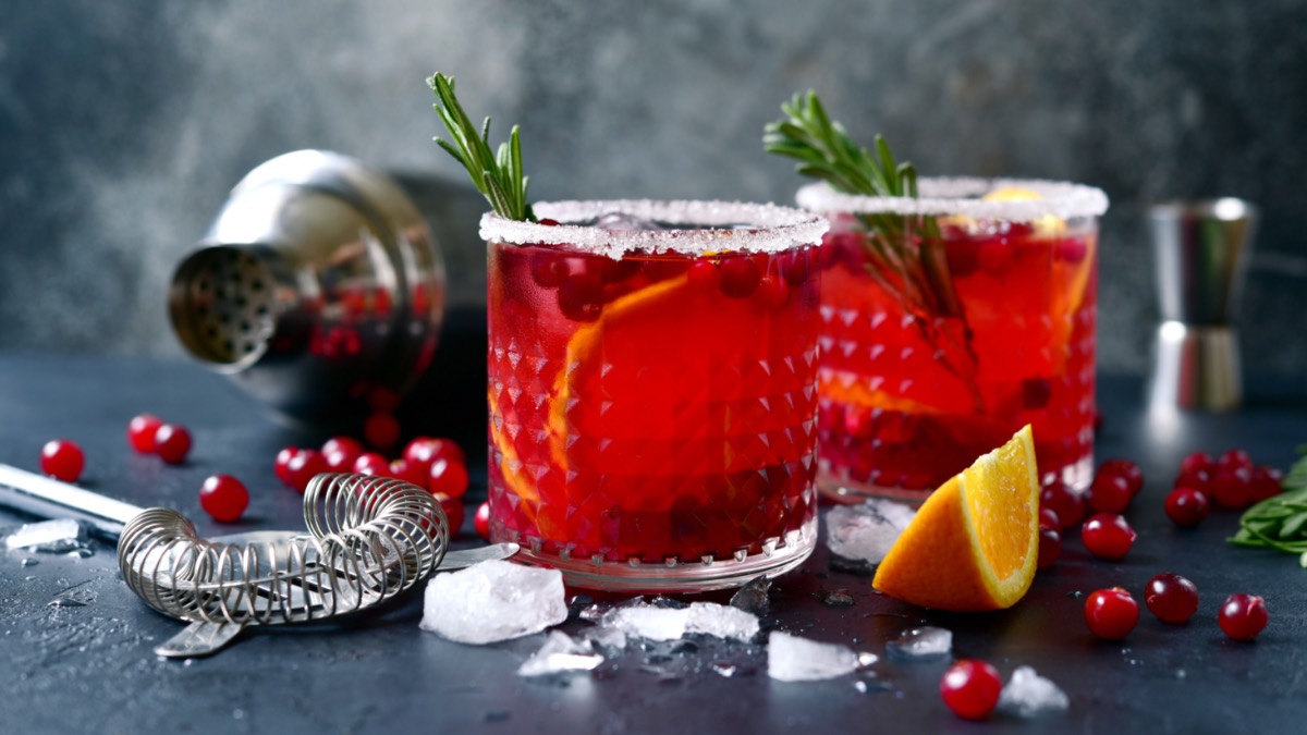 2 GlÃ¤ser Winter-Margarita mit Rosmarinzwigen. Im Hintergrund ein Cocktailshaker, EiswÃ¼rfel, Cranberrys, Orangen und Rosmarinzweige.