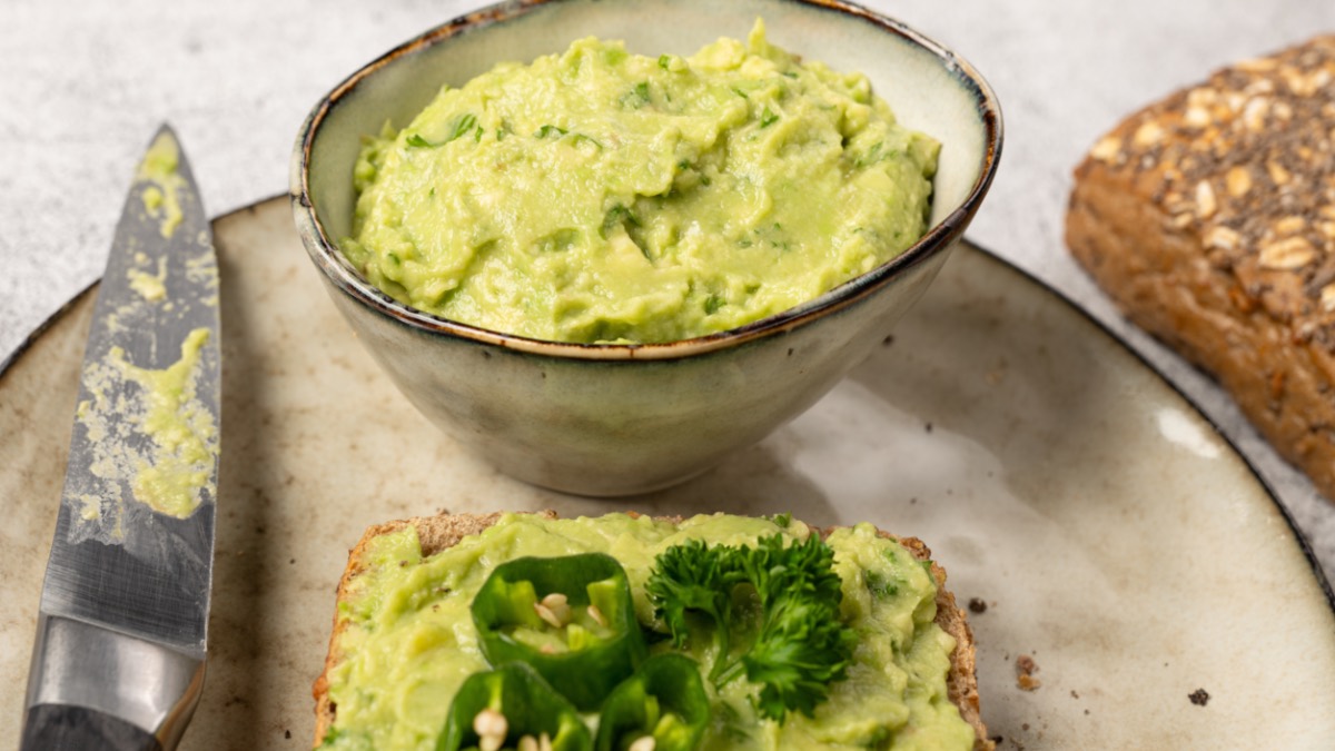 Ein groÃŸer Teller mit einer Schale Avocado-Cashew-Aufstrich, einer Scheibe Brot mit Aufstrich und einem Messer. Im Hintergrund liegt ein VollkornbrÃ¶tchen.