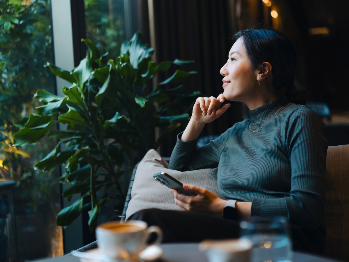 Junge asiatische Frau mit Smartphone, die aus dem Fenster schaut, wÃ¤hrend sie in einem CafÃ© sitzt und Kaffee trinkt.