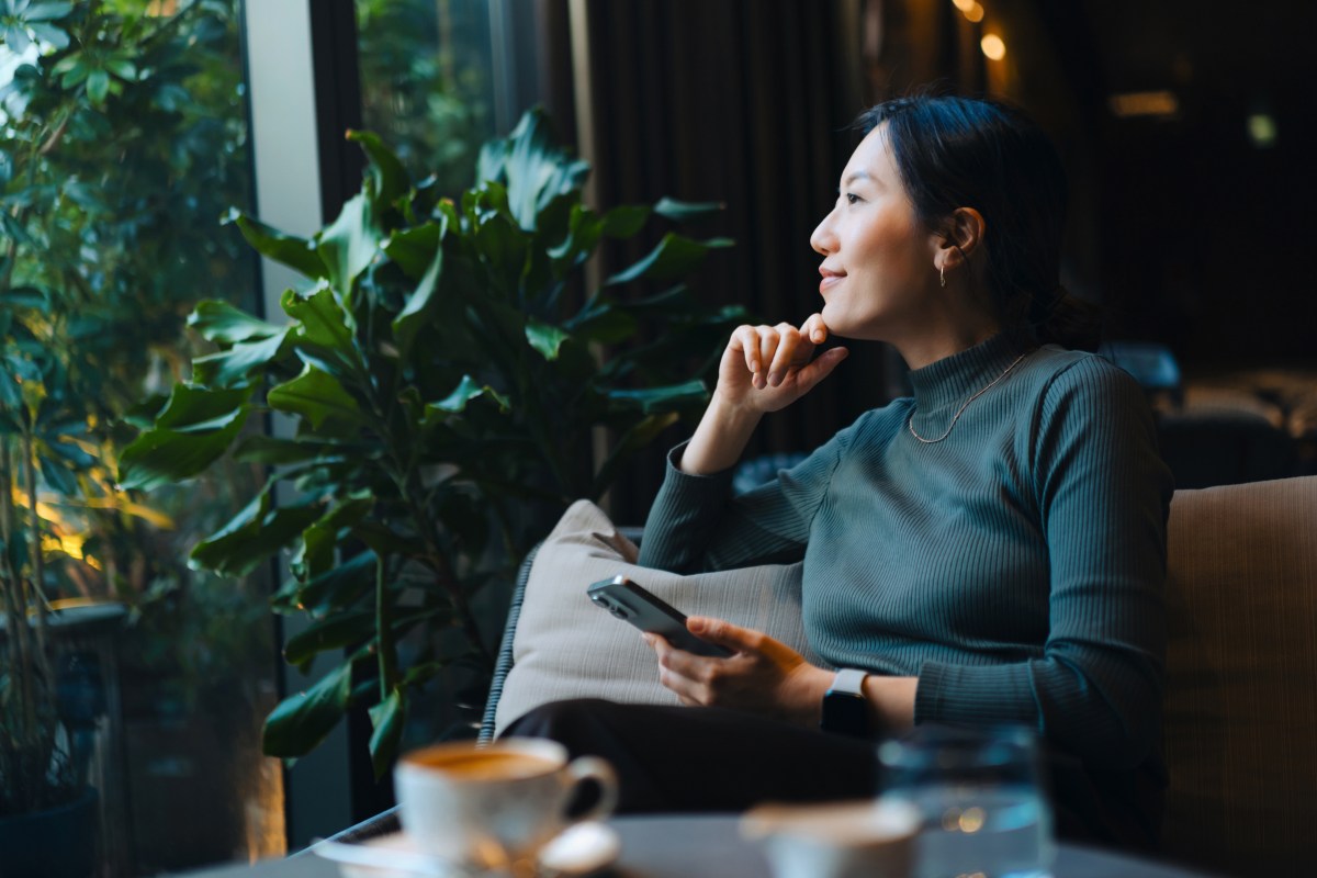 Junge asiatische Frau mit Smartphone, die aus dem Fenster schaut, wÃ¤hrend sie in einem CafÃ© sitzt und Kaffee trinkt.