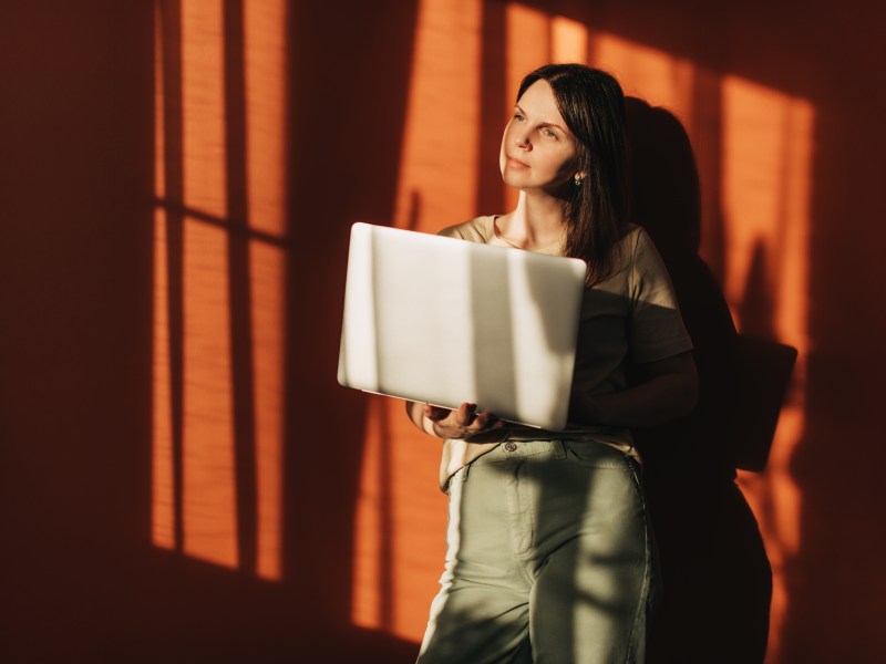 Eine Frau mit einem Laptop auf dem Hintergrund einer Terrakottawand, die Sonne scheint hell.