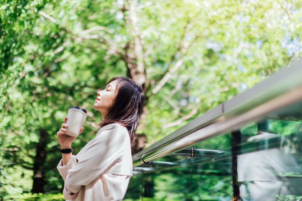 Asiatische Frau mit geschlossenen Augen, hält eine wiederverwendbare Kaffeetasse in der Hand und macht eine Pause im Park