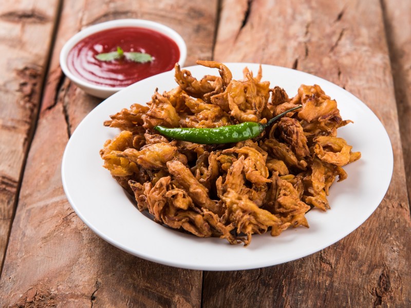 Indische GemÃ¼sekrapfen auf einem weiÃŸen Teller, dekoriert mit einer grÃ¼nen Chili und serviert mit einer roten SoÃŸe.