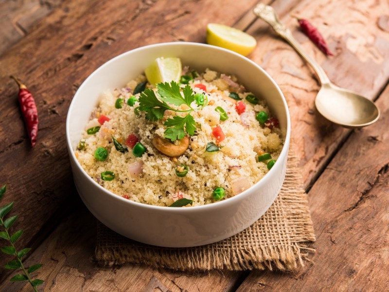 Eine Schale indisches Upma, HartweizengrieÃŸ, mit etwas GemÃ¼se und KrÃ¤utern, daneben ein LÃ¶ffel.