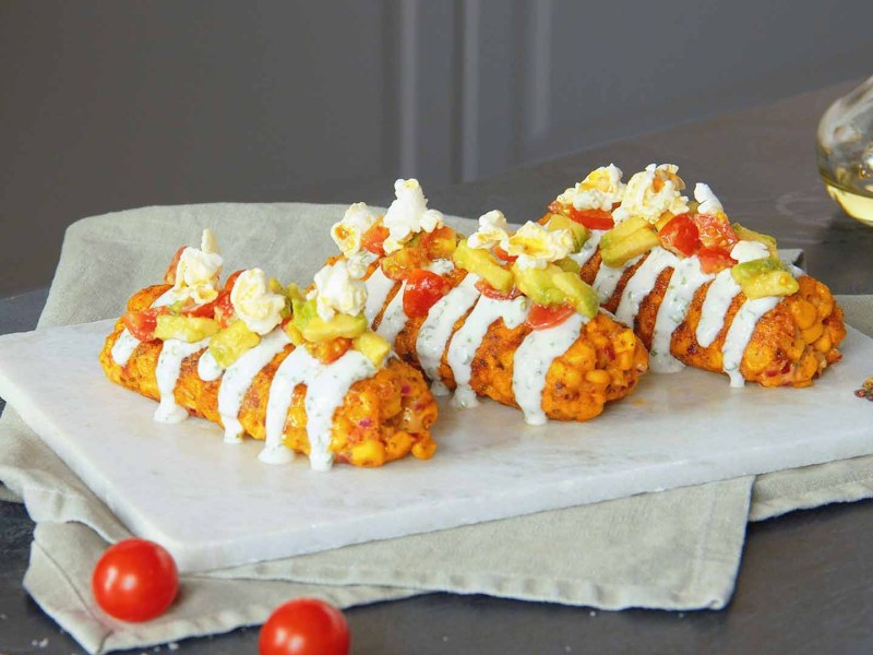 3 KÃ¤se-Mais-Kroketten auf einem Teller, garniert mit Sour Cream, Tomaten-Avocado-Salat und Popcorm.