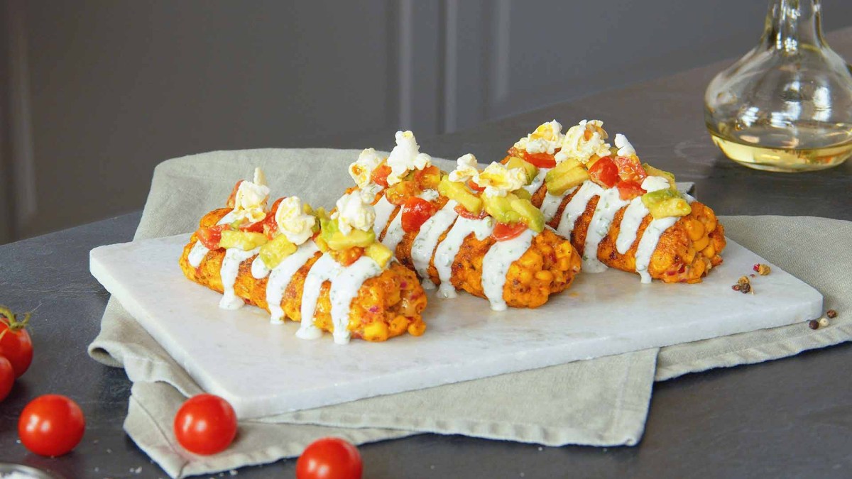 3 KÃ¤se-Mais-Kroketten auf einem Teller, garniert mit Sour Cream, Tomaten-Avocado-Salat und Popcorm.