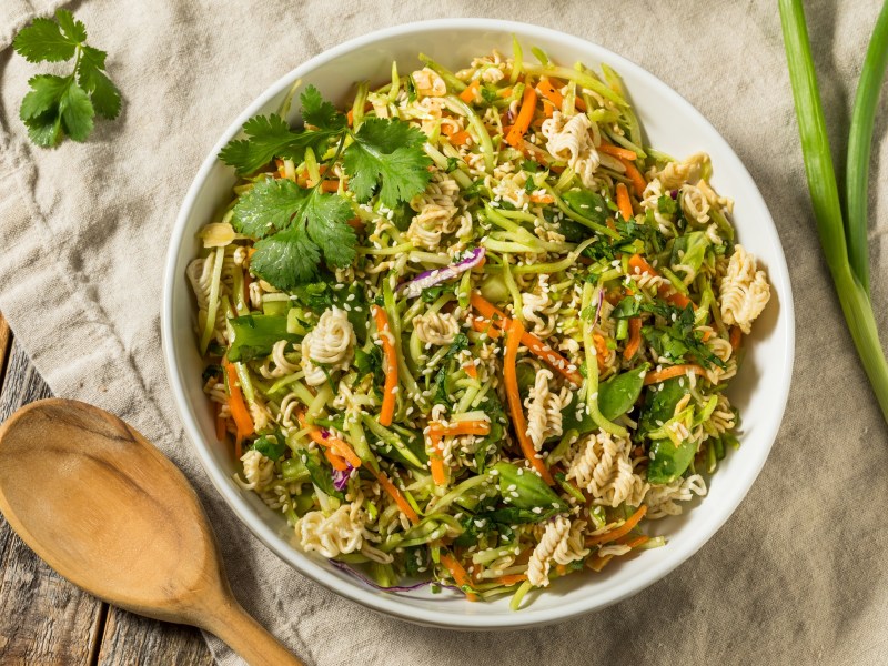 Knuspriger Ramen-Salat in einer weiÃŸen SchÃ¼ssel, dekoriert mit etwas frischem Koriander. Daneben liegen ein HolzlÃ¶ffel und eine Stange FrÃ¼hlingszwiebel.