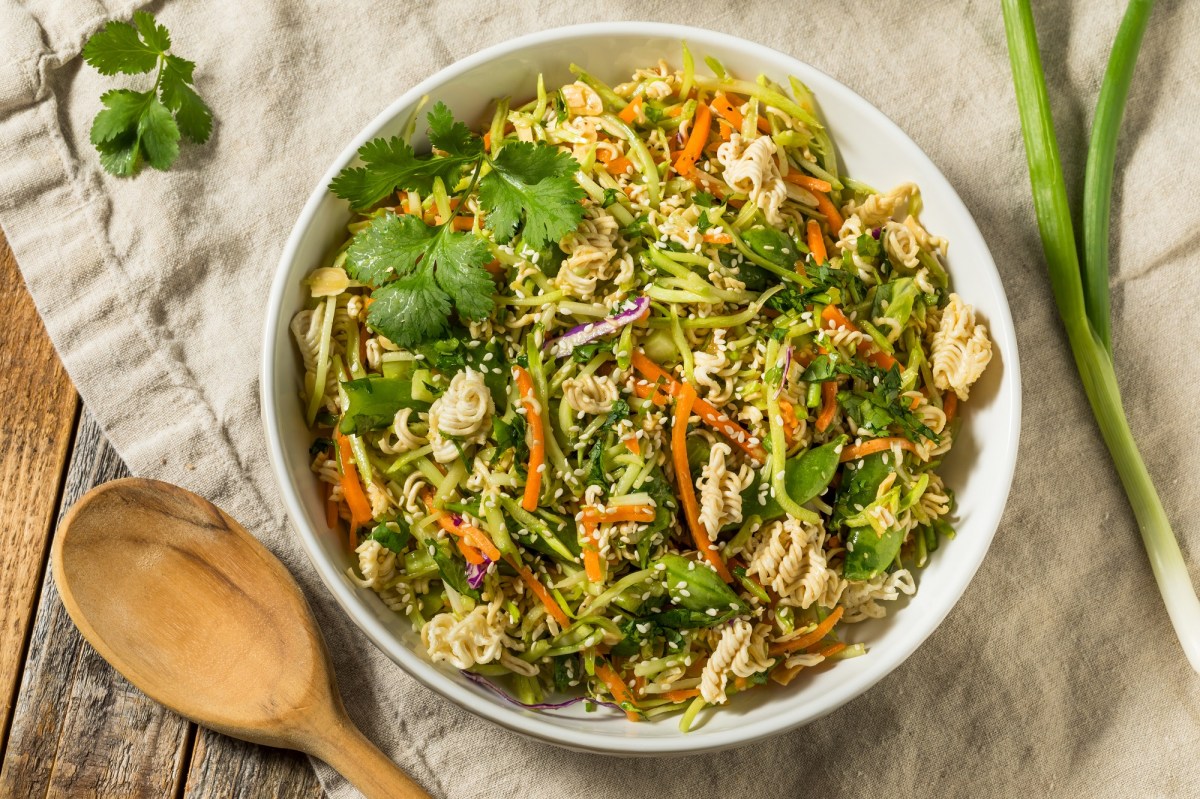 Knuspriger Ramen-Salat in einer weiÃŸen SchÃ¼ssel, dekoriert mit etwas frischem Koriander. Daneben liegen ein HolzlÃ¶ffel und eine Stange FrÃ¼hlingszwiebel.