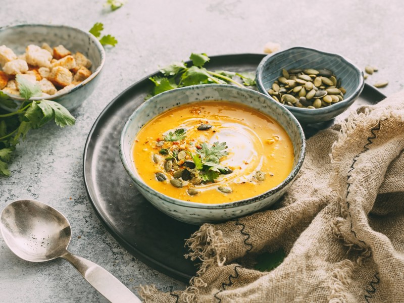 Eine SchÃ¼ssel KÃ¼rbis-Curry-Suppe mit Kernen, CroÃ»tons und KrÃ¤utern verfeinert, daneben je eine SchÃ¼ssel mit Kernen und CroÃ»tons.