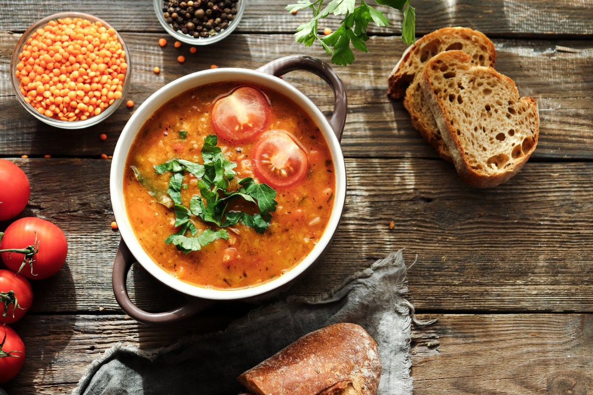 Eine SchÃ¼ssel mit Henkeln, darin Linsen-Tomaten-Suppe, garniert mit halbierten Tomaten und Koriander, daneben frische Tomaten, Brotscheiben, eine Schale Linsen, Draufsicht.