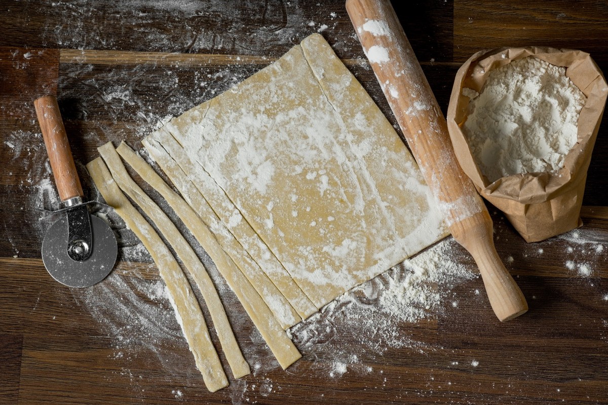 Nudelteig ohne Ei, ausgerollt und teilweise geschnitten, auf einer dunklen Holzunterlage. Daneben liegen ein Nudelholz und ein Pizzaschneider.