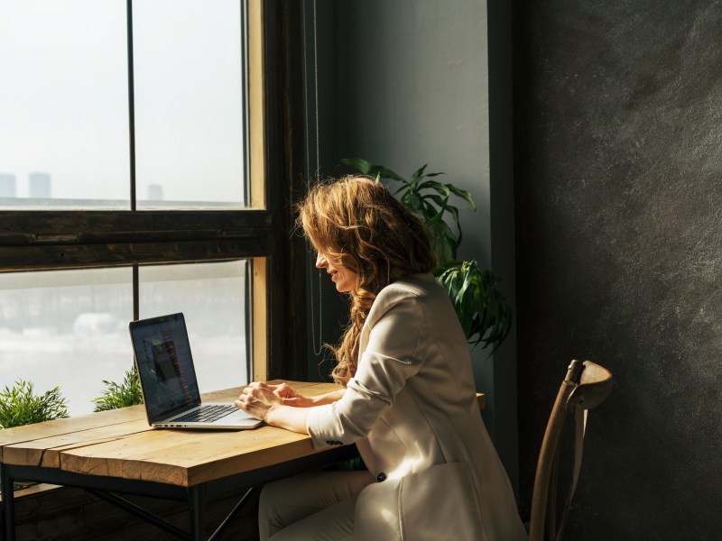 Eine blonde Frau mittleren Alters sitzt am Tisch vor einem Laptop am Fenster.