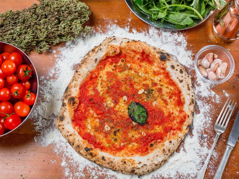 Pizza Marinara mit TomatensoÃŸe, Knoblauch und einem Blatt Basilikum in der Draufsicht, daneben eine Schale Tomaten, getrockneter Oregano, Basilikum und Besteck,