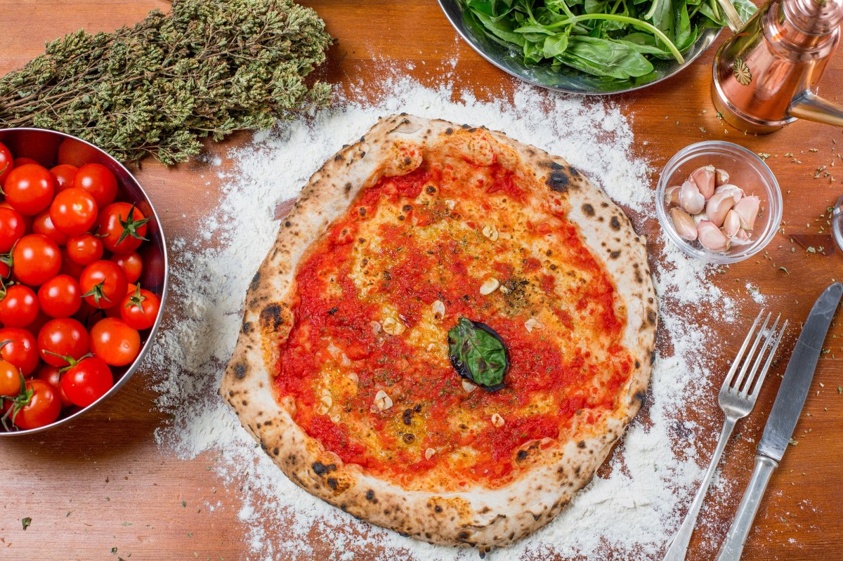 Pizza Marinara mit TomatensoÃŸe, Knoblauch und einem Blatt Basilikum in der Draufsicht, daneben eine Schale Tomaten, getrockneter Oregano, Basilikum und Besteck,