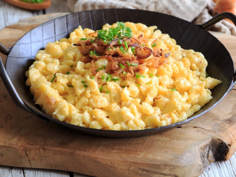 Eine gusseiserne Pfanne vegane KÃ¤sespÃ¤tzle mit RÃ¶stzwiebeln und Petersilie, Nahaufnahme.