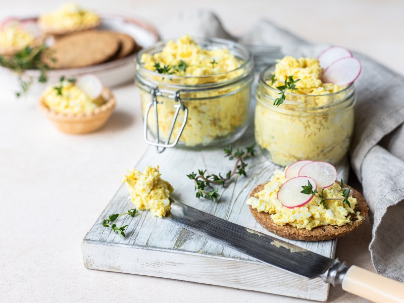 Zwei GlÃ¤ser veganer Eiersalat auf einem Brett, darauf auch eine runde Scheibe Schwarzbrot mit Eiersalat und ein Messer.