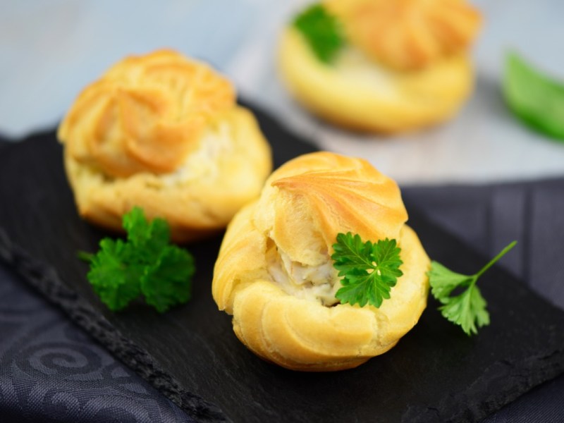 Drei Windbeutel mit FrischkÃ¤se-FÃ¼llung auf einer Schiefertafel. Garniert sind sie mit Petersilie.