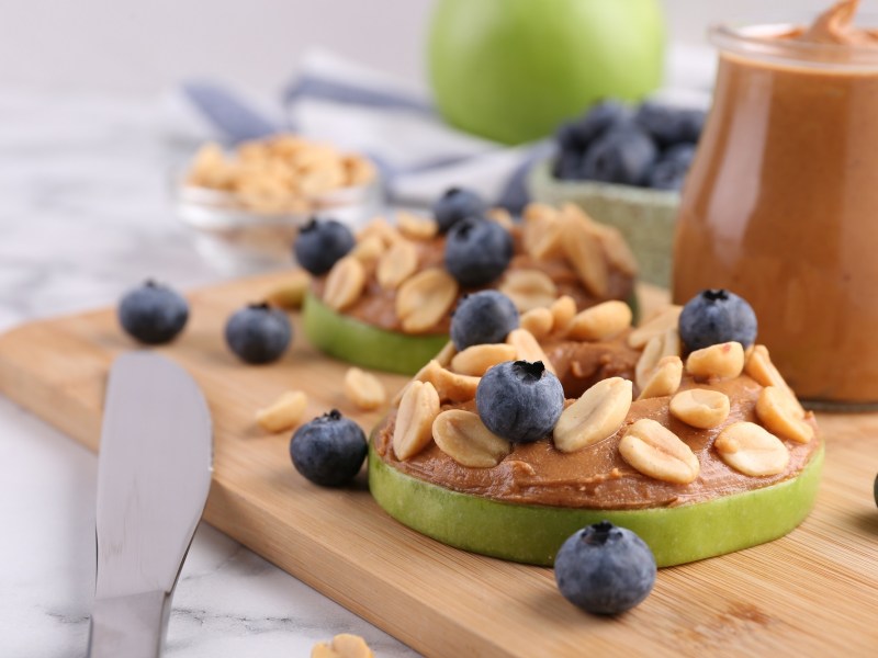 Apfel-Erdnussbutter-Bites auf einem Holzbrett. Sie sind mit Blaubeeren und ErdnÃ¼ssen garniert.