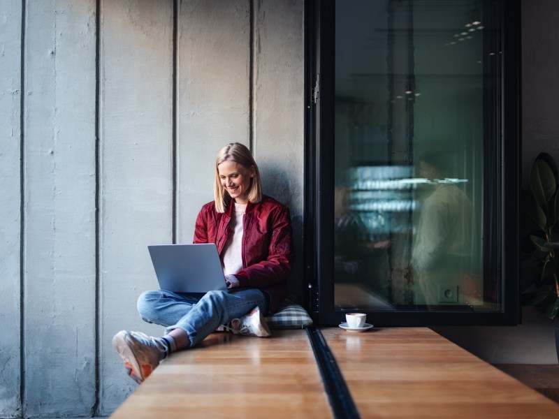 Eine junge Frau in einem lässigen Outfit lächelt, während sie ihren Laptop an einem gemütlichen Fensterplatz im Café benutzt.