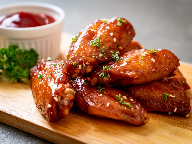 Ein paar Chicken Wings aus dem Airfryer aus einem Brett, bestreut mit Petersilie, daneben eine weiÃŸe Schale Ketchup.