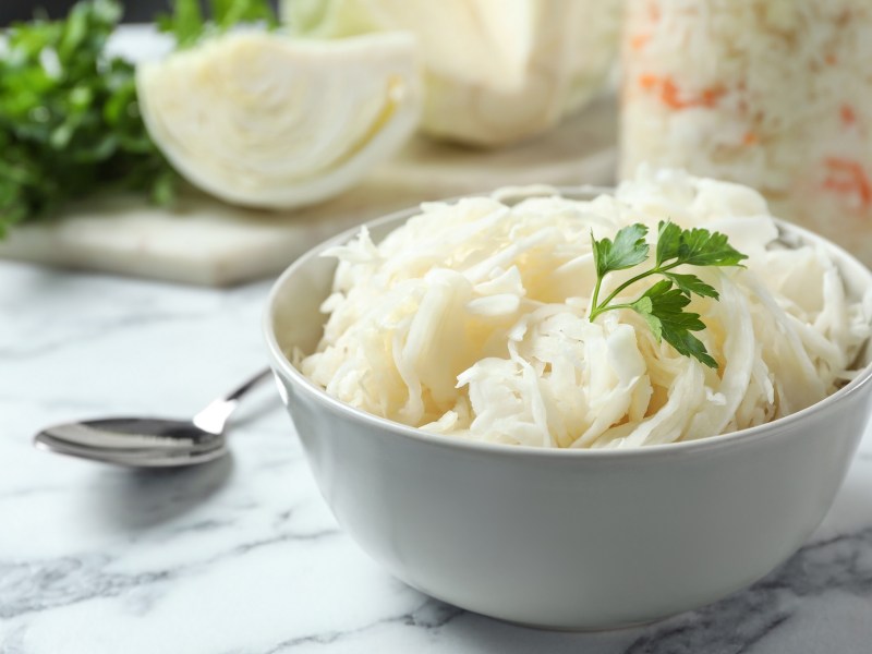 Eine SchÃ¼ssel sÃ¼dtiroler Krautsalat, daneben ein LÃ¶ffel, im Hintergrund ein Glas mit dem Salat und ein KohlstÃ¼ck.