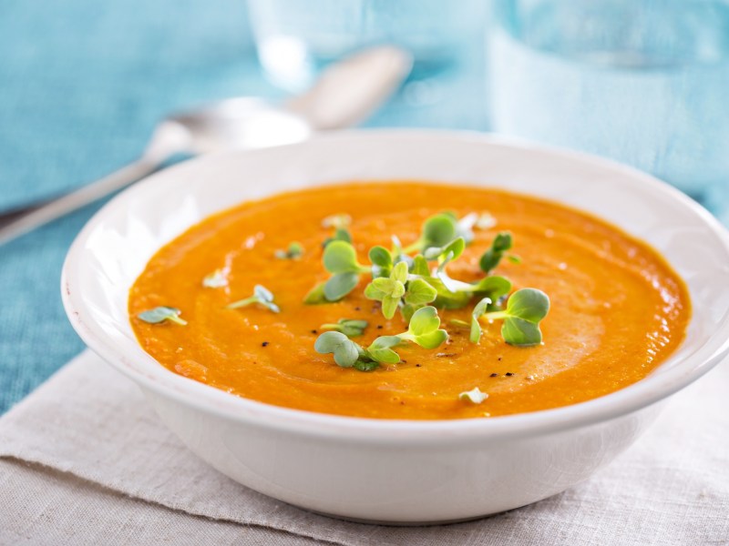 Ein tiefer Teller Zucchini-MÃ¶hren-Suppe garniert mit KrÃ¤utern auf einem Platzdeckchen, Dahinter unscharf ein Glas Wasser, Nahaufnahme.