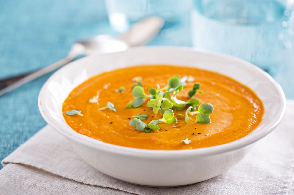 Ein tiefer Teller Zucchini-MÃ¶hren-Suppe garniert mit KrÃ¤utern auf einem Platzdeckchen, Dahinter unscharf ein Glas Wasser, Nahaufnahme.