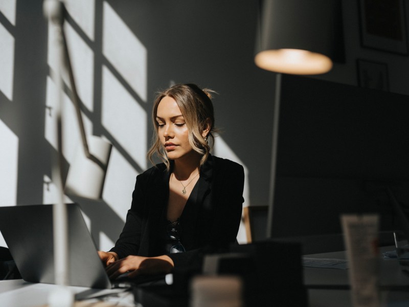 Eine junge Frau mit blonden Haaren sitzt im Büro und arbeitet am Laptop.